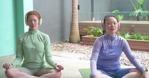Two women meditating on patio wearing headphones and athletic zip tops