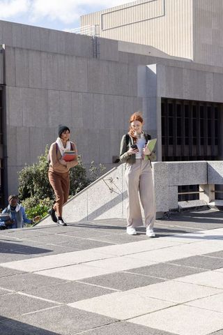 Redhead student checking phone while walking with diverse classmate on urban campus plaza