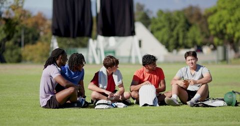 Young diverse male teammates sitting on grass sharing snacks and laughing after game