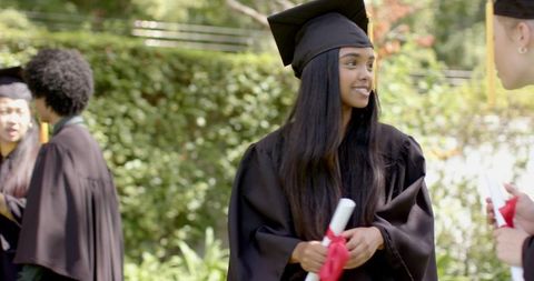 Outdoor graduation celebration featuring female graduate holding diploma and conversing