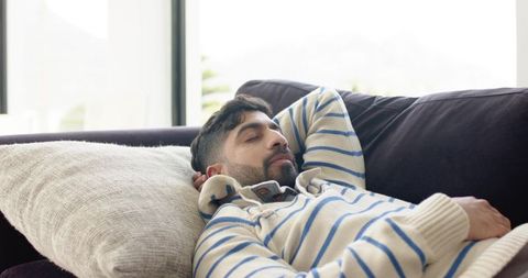 Indian man reclining on modern sofa in living room wearing cream blue striped sweater