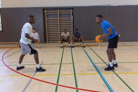 Two players running one-on-one basketball drill in indoor gym as diverse teammates watch