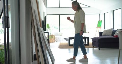 Senior woman walking through bright minimalist studio past stacked canvases and easel