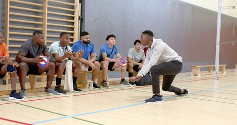 Coach demonstrating forearm pass to diverse male volleyball team on indoor bench during practice