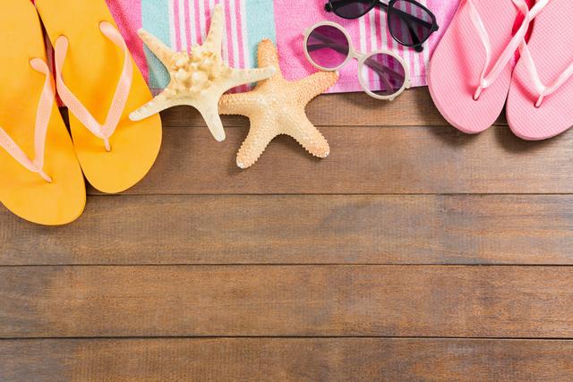 Perfect for travel and vacation-themed use, this image captures a variety of beach essentials such as flip flops, sunglasses, a towel, and starfish on a wooden board. Ideal for marketing summer products, travel brochures, and holiday promotions.