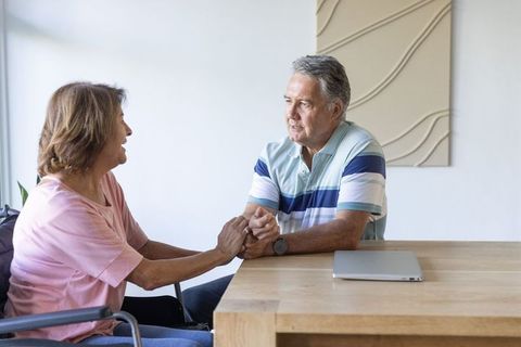 Mature man and senior woman in wheelchair holding hands and sharing tender conversation at table