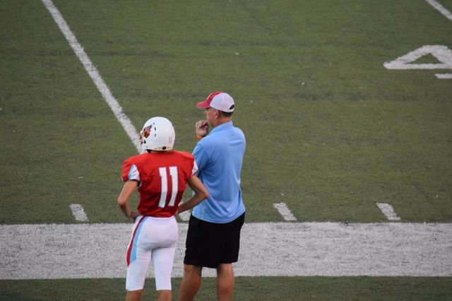 Coach in blue shirt and player in red jersey standing on the sideline of a football field, focused on game strategy. Useful for sports articles, teamwork concepts, and mentorship visuals, emphasizing leadership and coaching roles in athletics.