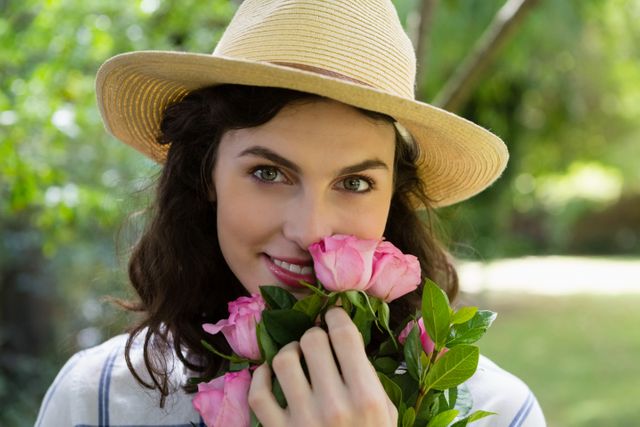 Woman wearing a hat and holding pink roses while smiling in a garden. Ideal for use in lifestyle blogs, gardening websites, floral advertisements, and summer-themed promotions.