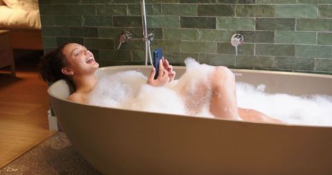 Laughing african american woman relaxing in bubble bath holding smartphone in bathroom