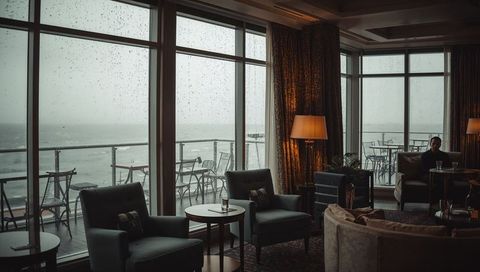 Sitting man in moody seaside lounge watching rain through wet floor-to-ceiling windows