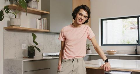 Young man leaning on modern kitchen island wearing pink tee, pearl necklace, smartwatch