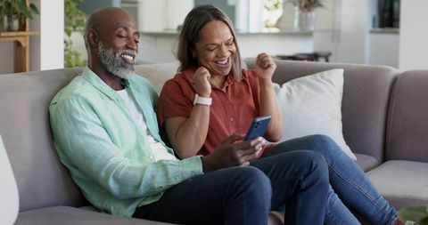 Mature couple celebrating together on sofa while viewing smartphone and wearing smartwatch