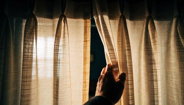 Hand pulling sheer curtain revealing soft morning light through pleated woven beige fabric