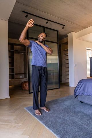 Mature african man stretching shoulder in modern minimalist bedroom, home wellness routine