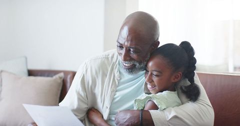 African american grandfather hugging granddaughter on couch while reading letter, smiling