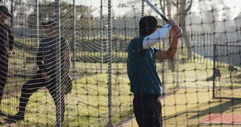 Male batter practicing swing inside netted batting cage at outdoor baseball field