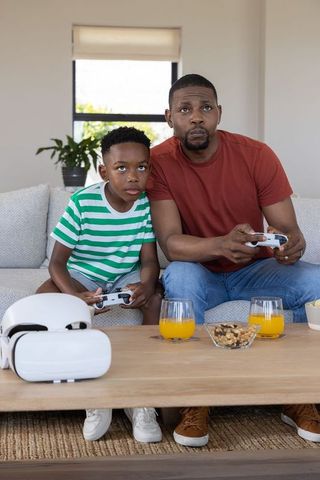 African American father and son gaming together on couch holding white controllers