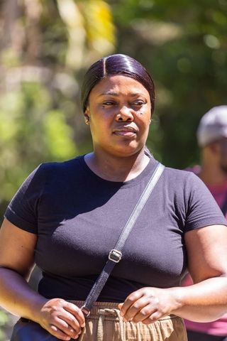 African woman wearing black tee and crossbody bag standing in sunlit park expressing calm