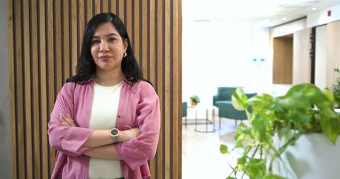 Confident professional woman standing arms crossed in modern office lounge and greenery