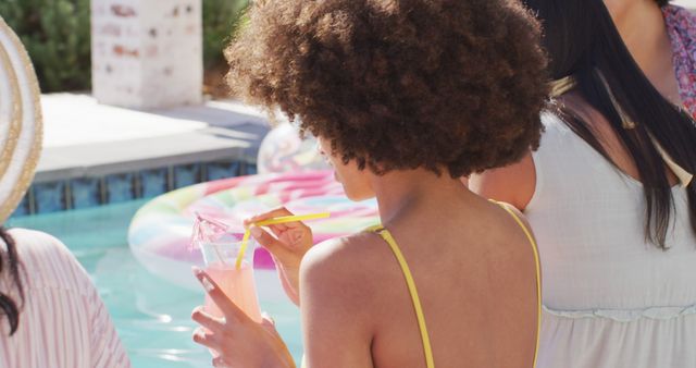 Young woman with afro hair sipping a drink from a straw while standing near a pool. Ideal for summer vacation themes, relaxation, leisure activities, and tropical getaways. Perfect for promotions related to holiday packages, beverage advertisements, and lifestyle blogs.