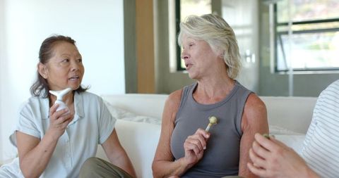 Mature diverse women chatting on sofa holding skincare tools and cup during self-care at-home