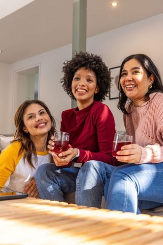 Diverse friends laughing and holding drinks on couch during cozy home gathering