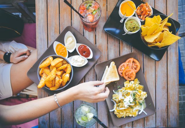 Group of friends enjoying a casual brunch, sharing various appetizers such as dipping sauces, seasoned potatoes, nachos, and a salad. Overhead perspective showcases colorful food and drinks. Ideal for ads, social media, food blogs, or promoting restaurants and culinary services.