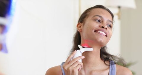 Smiling woman using red light therapy device on neck at home for glowing skin and selfcare