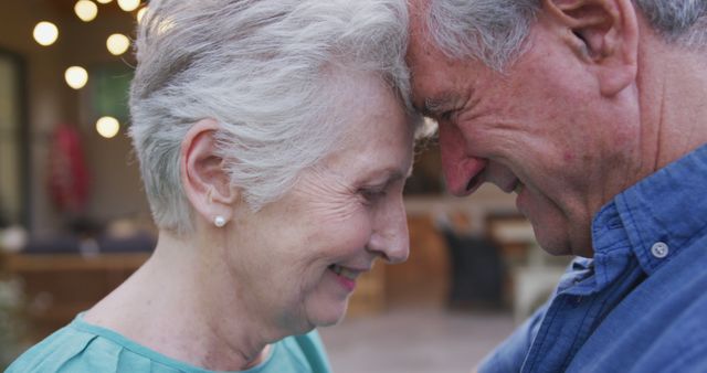 Elderly couple sharing a tender, intimate moment outdoors. Both individuals are in close proximity, resting their foreheads together and smiling, showcasing their deep bond and affection. Ideal for use in advertisements for retirement communities, senior care services, or products aimed at the elderly. It can also be utilized in campaigns promoting love and relationships in later stages of life.