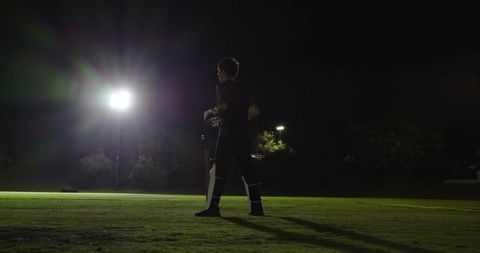 Young cricket batsman standing under floodlights holding bat wearing pads at night
