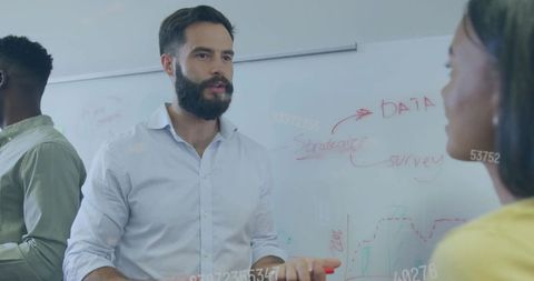 Bearded leader pointing to data on glass board while colleague listens during brainstorm