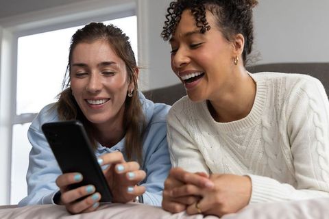 Multiracial friends laughing while scrolling smartphone in cozy bedroom, candid lifestyle