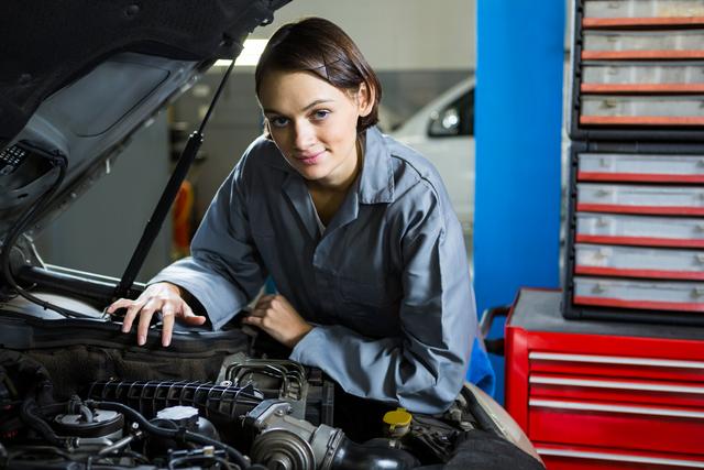 Young female mechanic servicing a car in a modern repair garage. She is wearing a work uniform and is surrounded by various tools and equipment. This image can be used for promoting automotive services, illustrating female empowerment in skilled trades, or showcasing professional car repair services.