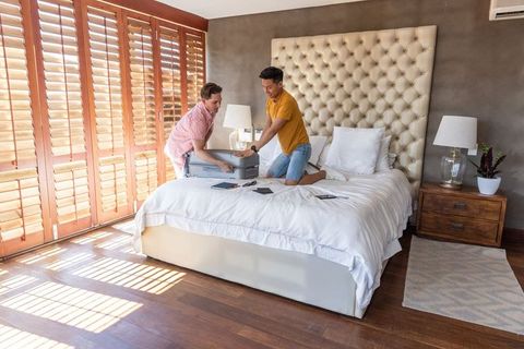 Sunlit master bedroom friends packing silver hard-shell suitcase on bed preparing travel