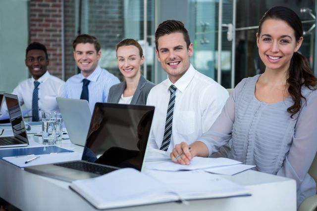Diverse group of business professionals sitting in a conference room, smiling at the camera. They are dressed in business attire and have laptops and documents in front of them. Ideal for use in corporate websites, business presentations, team collaboration concepts, and professional development materials.