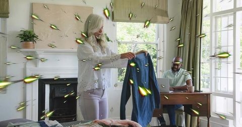 Senior white woman holding shirt while black man working at laptop in sunlit home office
