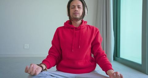 Young man meditating cross-legged in red hoodie with headphones by sunlit window