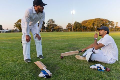 Diverse teammates practicing cricket at dusk on grassy field wearing white kits and pads