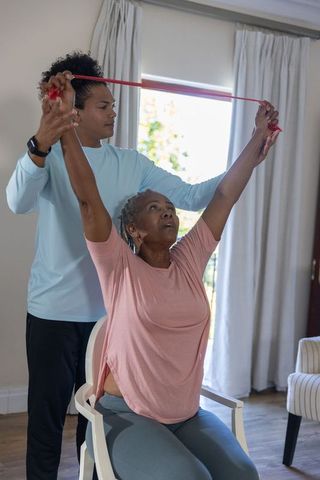 African american senior doing seated overhead resistance band exercise with caregiver