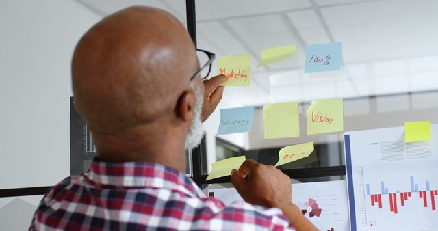 Bald businessman placing sticky notes with important tasks on a transparent board in a modern office. This image highlights concepts of planning, organization, and productivity. Suitable for content related to business strategy, project management, professional development, and time management.