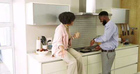 Diverse couple cooking together in sunlit modern kitchen, sipping coffee and laughing