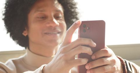 Mid-adult african american man using red smartphone by sunlit window, relaxed candid shot
