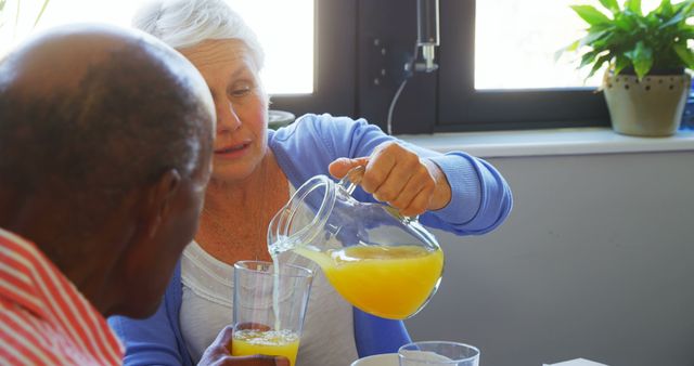 Senior woman pouring fresh orange juice into glass for her elderly friend while sitting at table. Image depicts warm hospitality, health-conscious lifestyle, and strong companionship in senior years. Suitable for promoting elderly care services, healthy living, or items related to senior activities.