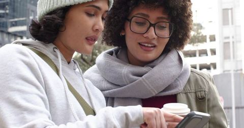 Friends Checking Phone Together on Urban Sidewalk Holding Coffee, Wearing Scarf and Beanie