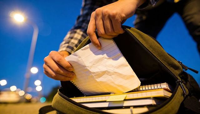 Student removing crumpled notebook paper from backpack on roadside at dusk