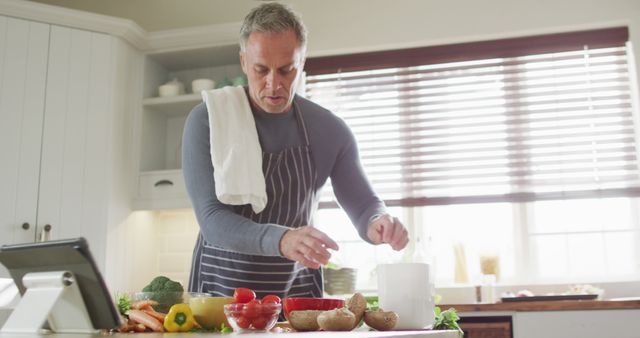 Mature man preparing a meal in a modern kitchen. He is chopping vegetables and using a tablet for recipe guidance. This can be used for content related to healthy living, home cooking, recipe blogs, or promotional materials for kitchen products and technology integration in cooking.