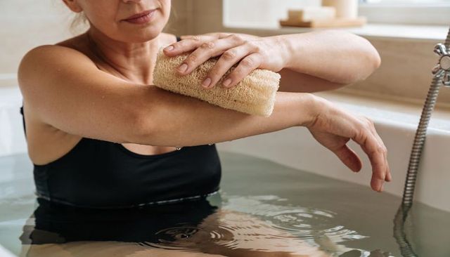 Middle-aged woman exfoliating forearm in bathtub with natural loofah, home spa relaxation