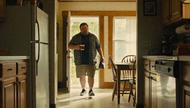 Middle-aged man entering kitchen carrying coffee mug and small bag, plaid shirt, morning light