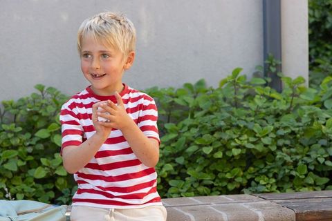 Smiling boy clapping hands on brick ledge in garden with backpack nearby
