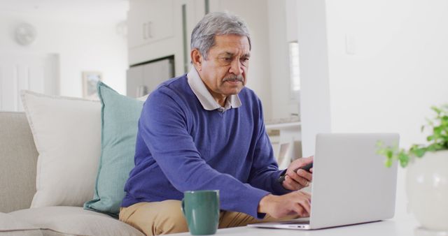 Image of senior biracial man using smartphone and laptop sitting in living room, smiling. Retirement, communication, inclusivity and senior lifestyle concept.